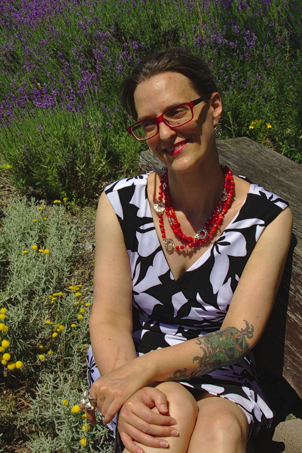 Photo of a the author sitting in a field wearing a black and white floral dress and a red beaded necklace