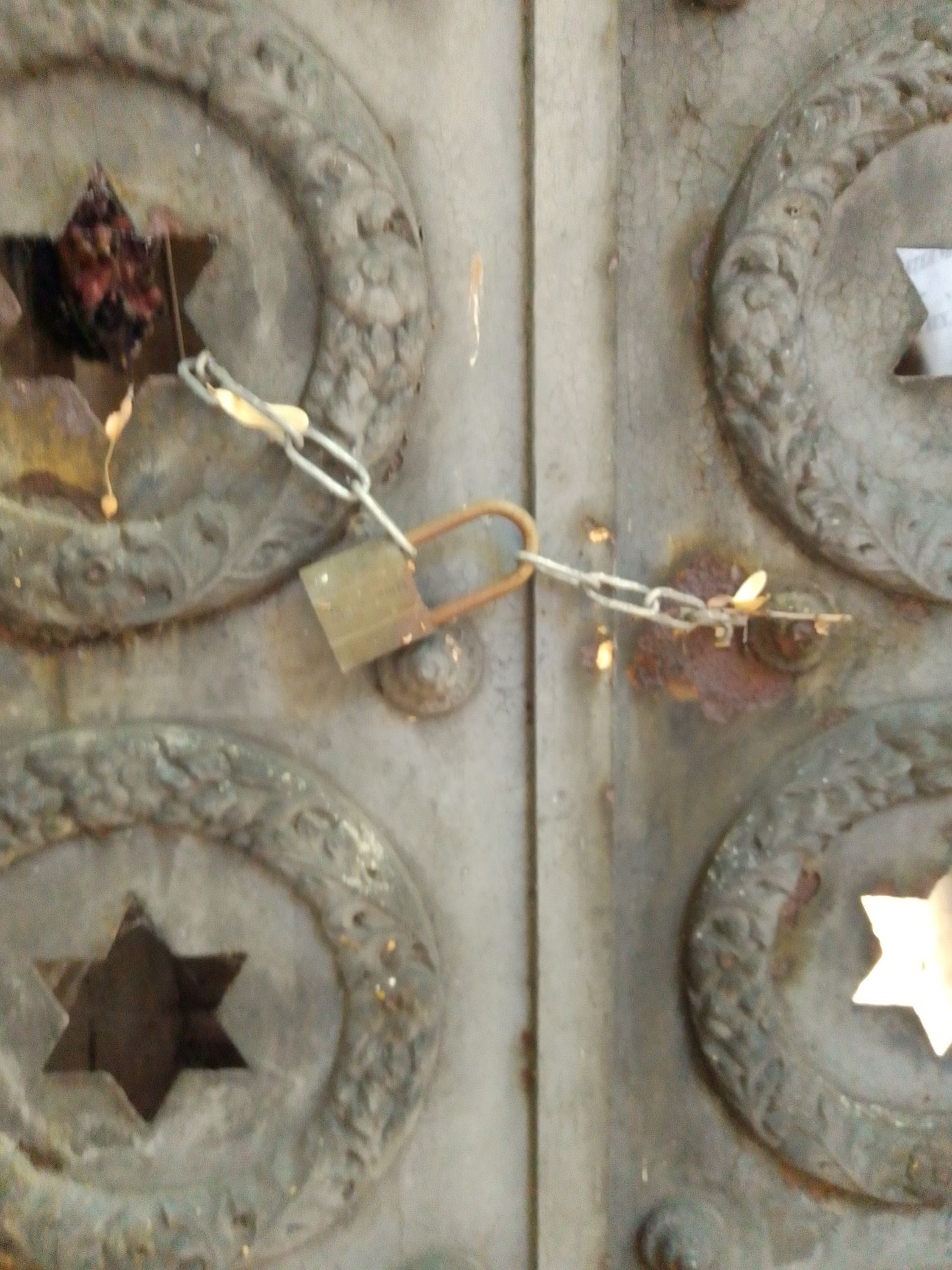 Close up of a crypt door with star shaped opening inside raised circles. The door is locked with a thin chain closed by a small padlock.