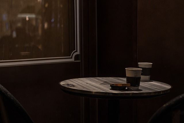 Two coffee cups sit on a wooden table in front of a window in a dark room